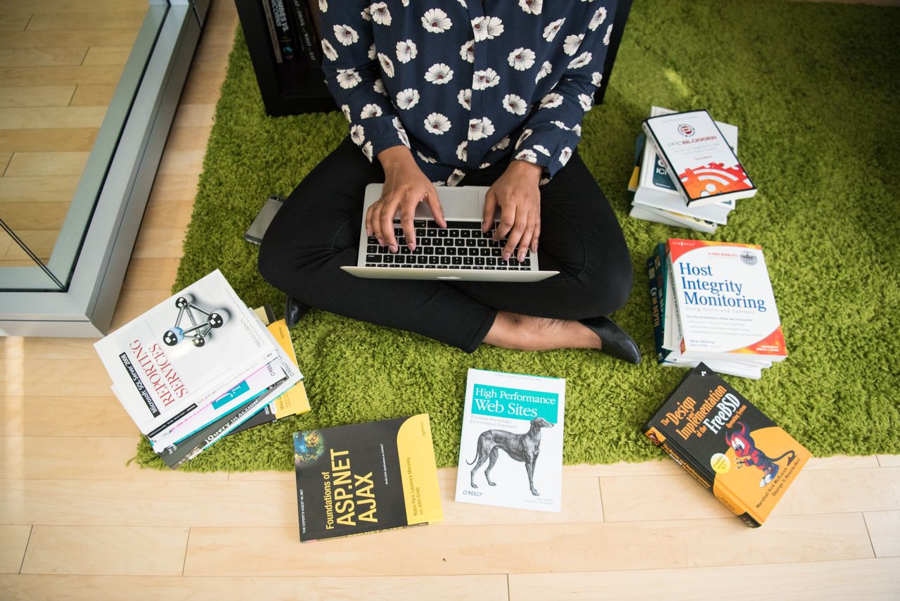 Services-01 Woman sitting on green rug working on laptop, surrounded by technology books in a modern room.